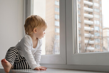 Fototapeta na wymiar Cute caucasian blonde baby girl about 1,2 years old sitting on sill looking through window at street autumn view.Infant,toddler,kid safety,secuity.