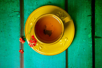 Yellow cup with black aromatic tea inside, red spindle shrub flower on saucer view from above. Mug stands on wooden green table top view. Beautiful composition flatly with place for text. Hot beverage