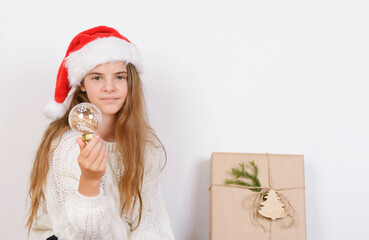 Young girl in white sweater and red santa hat holds bauble christmas decorations direct look