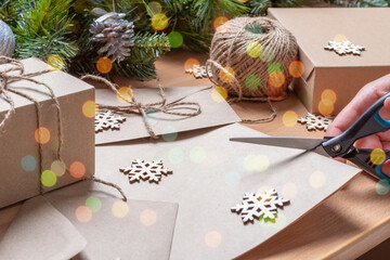 hand cutting recycled paper with scissors on the table with artificial fir tree with cones, envelope, twine, and wooden snowflakes