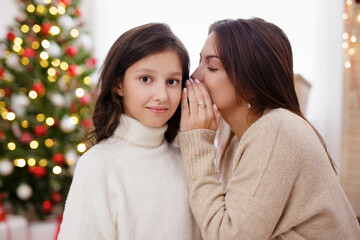 Christmas, family and motherhood concept - happy mother whispering secret to her daughter in decorated living room