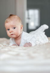 The child, lying on his stomach, raises his head and looks into the camera against the background of a bright interior. Vertical photography