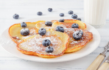 pancakes with blue blueberries and powdered sugar on a plate,  glass of milk on a white wooden