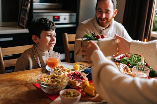 Close-up View From Back Of Loving Young Mother Giving Festive Box With Christmas Present To Son Sitting At Dinner Feast Table During Holiday Family Party, Selective Focus, Blurred Background.