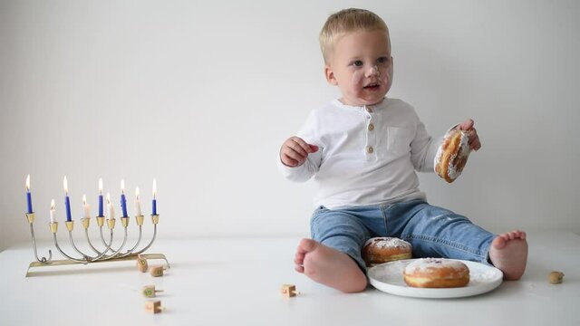 Kid in white t-shirt on white background celebrating Hanukkah. Jewish festival of lights. Child smile and happy. On table donuts and traditional menorah with lighting candles. Holiday in Israel 
