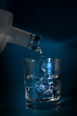 A transparent glass with ice and the neck of a bottle from which liquid is poured