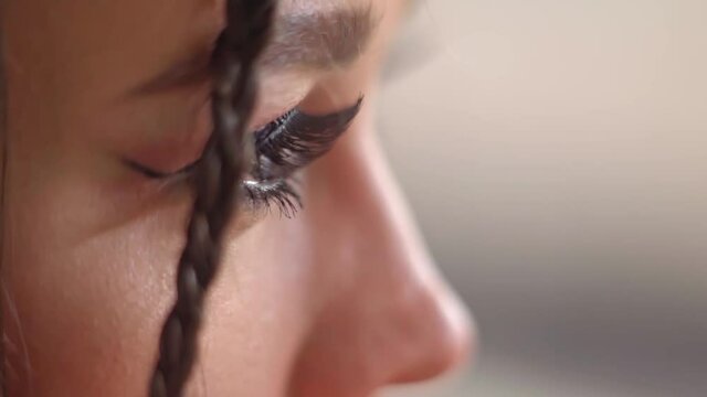 Close-up of the eyes with the long eyelashes of a pretty girl with beautiful makeup and braided African tail long hair turns on camera with a gleam of red light on her face. FHD video