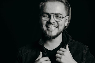 Black and white photo of young attractive guy laughing sincerely. Portrait of happy man with wide smile with teeth. Charismatic cute male with round glasses and modern stylish haircut. Studio shooting