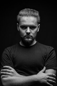 Serious guy in T-shirt and with beard in studio on black background and cross hands and staring in camera. Threatening look. Black and white photo, shadow under eyes adds negative emotion to model