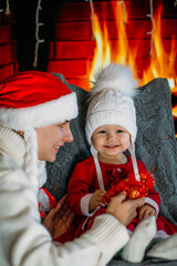 Fototapeta na wymiar Mom in a Christmas hat shows daughter in a red outfit festive decorations on the background of the fireplace
