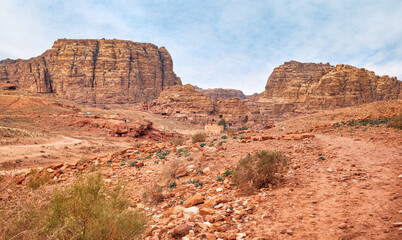 Fototapeta premium Typical landscape at Petra, Jordan, red dusty ground, mountains with carved buildings inside in distance