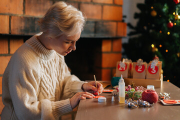Side view of happy young woman writing number on red bag by brush and white paint sitting at table. Attractive female making paper bags from kraft paper for advent calendar on Christmas Eve.