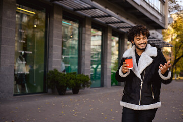 Young indian man in earphones talking while holding coffee to go in city 