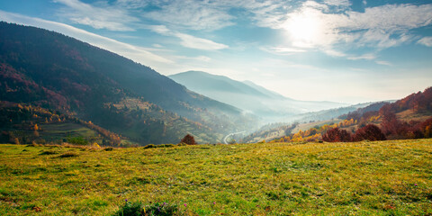 rural landscape at sunrise. beautiful autumnal mountain scenery. green grassy meadow on the hillside. fog down in the valley. sun and fluffy clouds above horizon