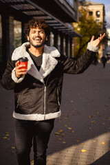 Positive indian man in earphones holding paper cup and singing on city street 