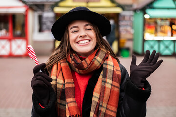 Young pretty smiling woman hold eat lollipop sweet striped candy celebrating open mouth white teeth. Cold weather, winter market holidays fair, travel resort. New Year Christmas decorations
