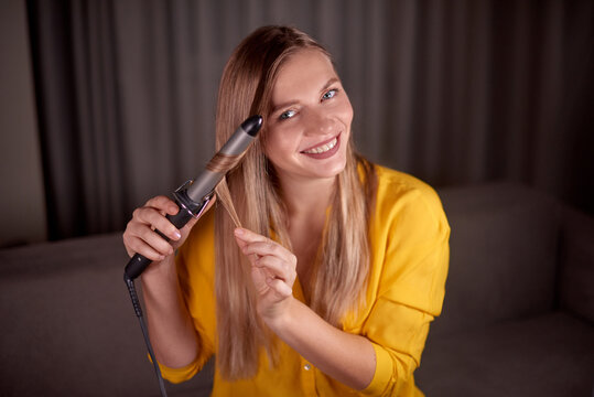attractive woman hairdresser sits looking directly at the camera and shows how to style hair with a curling iron