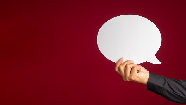 A speech bubble concept. Hand holding an empty white speech bubble over on a red background with copy space for text. Close-up photo