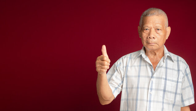 Portrait of a senior man showing thumbs up and looking at the camera with a smile while standing over a red background with space for text in the studio. Aged people and healthcare concept