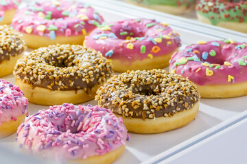 Round donuts in various glazes on the counter. Chocolate glazed donuts and pink glazed donuts. Various sprinkles with colored crumbs. Sale of confectionery. High-calorie and unhealthy foods