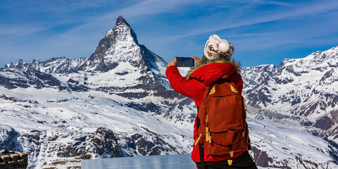Back view of young woman tourist in red cloth using smartphone taking apicture in Matterhorn...