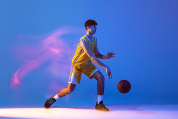 Naklejka premium Dynamic portrait of professional basketball player training with ball isolated on blue studio background in mixed neon light.