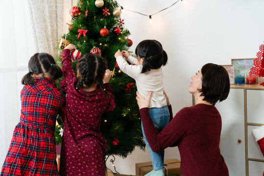 Rear View Of Happy Asian Mother Holding Up Her Baby Daughter While Her Other Kids Are Decorating Christmas Together In The Living Room At Home During The Day