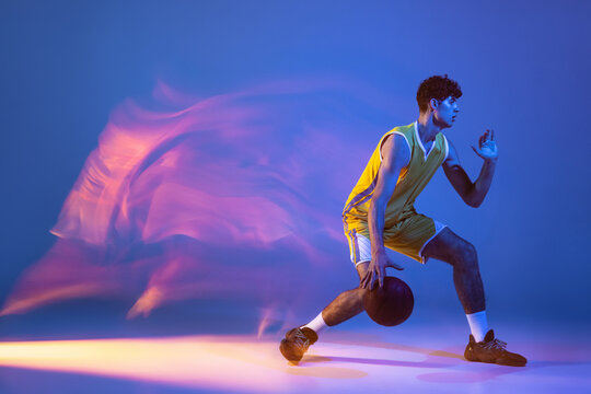 Dynamic portrait of professional basketball player training with ball isolated on blue studio background in mixed neon light.