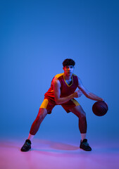 Naklejka na ściany i meble Studio shot of young man, professional basketball player playing basketball isolated on blue background in neon light.