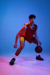 Fototapeta na wymiar Studio shot of young man, professional basketball player playing basketball isolated on blue background in neon light.