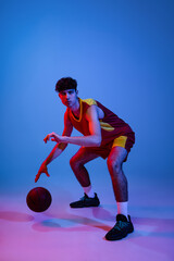 Studio shot of young man, professional basketball player playing basketball isolated on blue background in neon light.