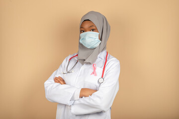 Portrait of an African American female doctor wearing a surgical mask, scarf and white coat posing with her arms crossed.