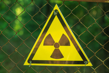 Close-up of a yellow triangular radiation sign on a wire mesh. In the background is a green forest....