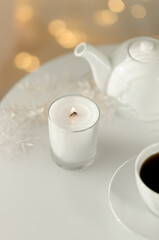Obraz na płótnie Canvas A white candle with a decorative white branch is burning on a white table with a cup of tea and a teapot. Candle flame and blurred lights
