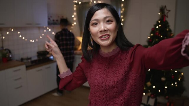 mobile cam view asian Japanese woman waving hi and gesturing boyfriend to come as they are preparing for Christmas dinner and having video call with friend at home