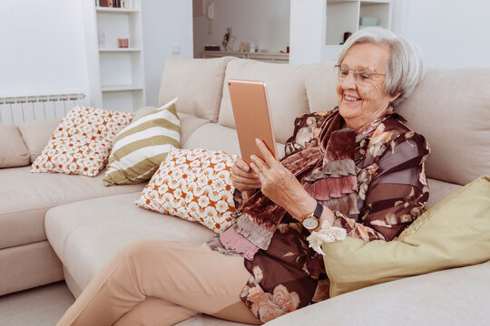 Happy 90s Older Mature Adult Woman Holding Digital Tablet Computer Conference Calling By Social Distance Virtual Family Online Chat Meeting Or Watching Video Sitting On Couch At Home.