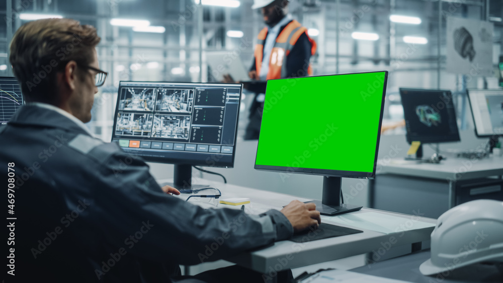 Fototapete Vehicle Factory Line Operator Working at Desk with Computer with Green Screen Display. In the Background African American Engineer Using Tablet and Looking at a Car Assembly Plant. 