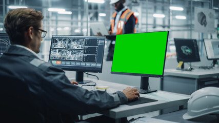 Vehicle Factory Line Operator Working at Desk with Computer with Green Screen Display. In the Background African American Engineer Using Tablet and Looking at a Car Assembly Plant.  - Powered by Adobe