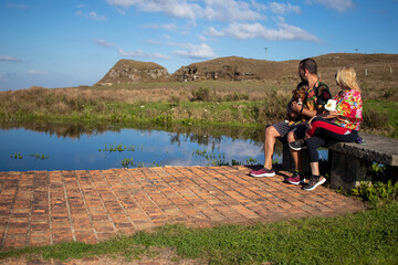 couple sitting by the lake, looking at the landscape, man holding a little black dog, sunny day with blue sky, photo view from a distance with space for text.