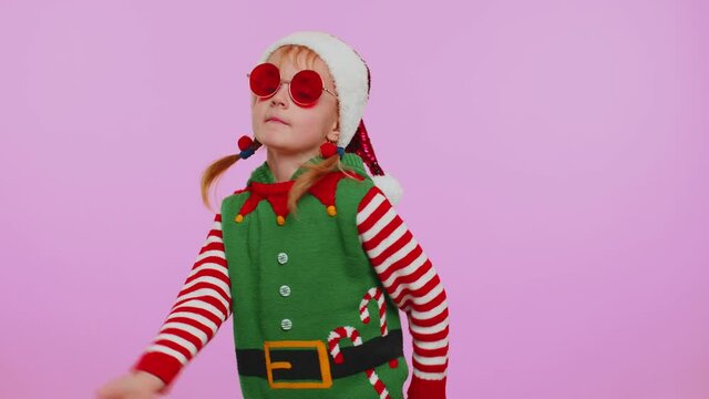 Toddler girl in Christmas Santa Claus helper Elf costume listening music dancing fooling around having fun expressive gesticulating hands isolated on violet background. Happy New Year merry holiday