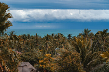 The view from the balcony to the rooftops, forest and palm leaves. In the background turquoise sea and blue cloudy sky