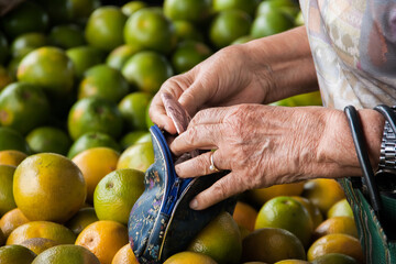 Mãos de uma pessoa idosa colocando dinheiro na bolsa em uma banca de laranjas.