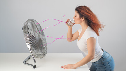 A red-haired Caucasian woman chills by the electric fan and drinks a cold drink. Climate control in...