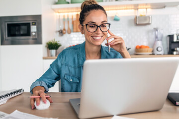 Smiling business woman working with laptop while talking with smartphone in the kitchen at home.