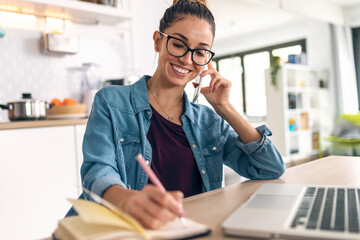 Smiling business woman working with laptop while talking with smartphone in the kitchen at home.