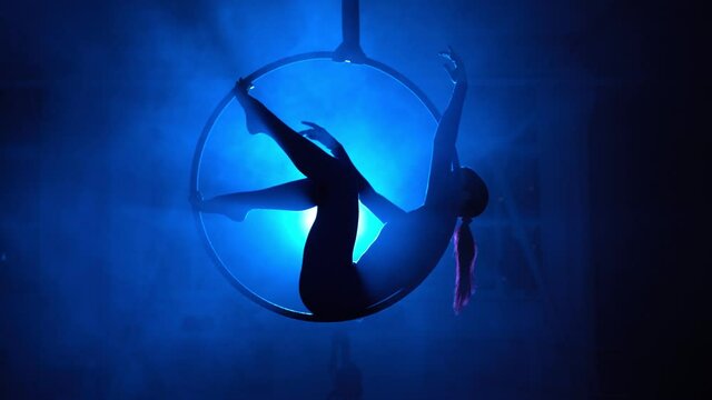 Silhouette, woman aerial gymnast performs a trick in the ring in a smoky room with backlit blue light. Neon lighting. 