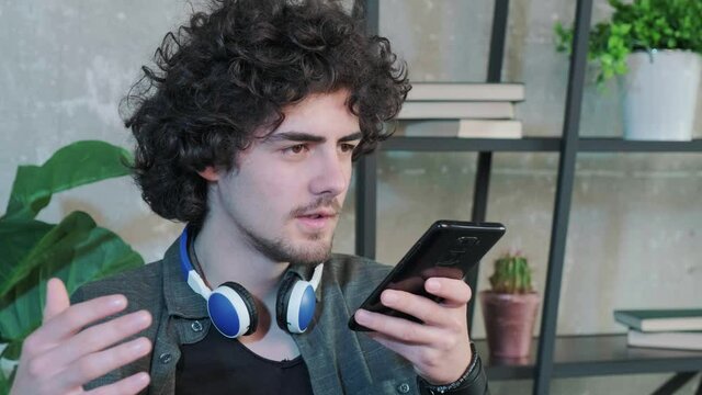 Young Caucasian man sit at desk at home office and talk on loudspeaker on smartphone. Millennial male use cellphone to record voice message.