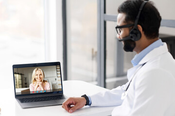 Indian male MD wearing headset, eyeglasses and medical gown giving online consultation to female...