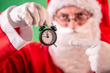 Santa Claus in red coat, red hat and round glasses, holding a small black alarm clock and pointing...