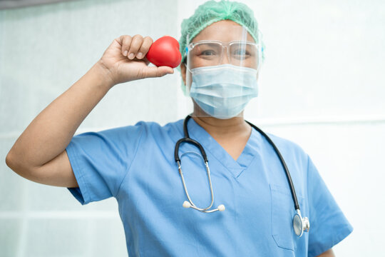 Asian Doctor Wearing Face Shield And PPE Suit New Normal To Protect Safety Infection Covid-19 Coronavirus Holding Red Heart In Hand At Hospital.